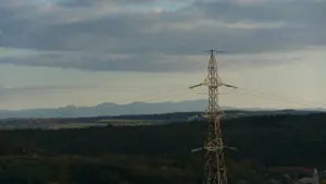 Aukštos įtampos elektros perdavimo linija rudenėjančiame peizaže prieš debesuotą dangų.
