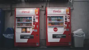 Dvieji „Coca-Cola“ gėrimų automatai su šiukšliadėžėmis šalia pastato sienos