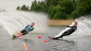Du jauni vandenlenčių sportininkai važiuoja vandens slidėmis ežere, keldami didelius vandens purslus ir bangas.