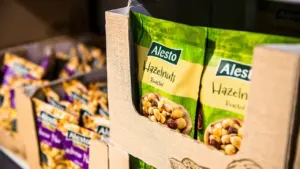 „Alesto“ prekės ženklo skrudintų lazdyno riešutų pakuotės sudėtos į kartoninę lentyną parduotuvės skyriuje.