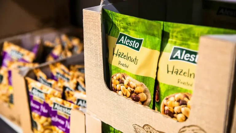 „Alesto“ prekės ženklo skrudintų lazdyno riešutų pakuotės sudėtos į kartoninę lentyną parduotuvės skyriuje.