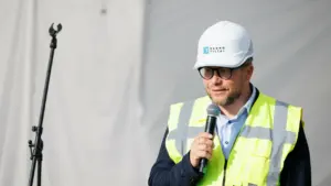 Vyras su baltu apsauginiu šalmu ir ryškia liemene kalba į mikrofoną statybvietės fone.