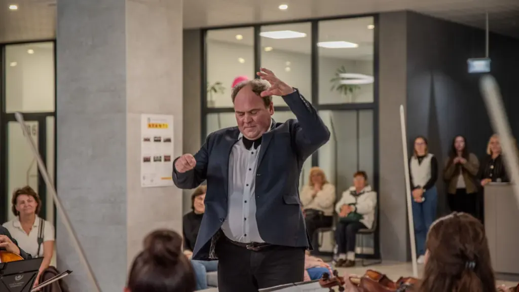 Dirigentas su tamsiu švarku energingai gestikuliuoja priešais muzikantus erdvioje šiuolaikiškoje koncertų salėje.