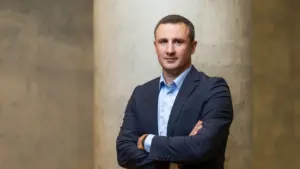 Trumpiantas vyras tamsiu švarku ir šviesiais marškiniais stovi sukryžiuotomis rankomis prie betono kolonos.