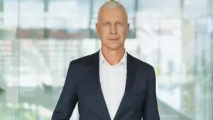 Šviesiaplaukis vyras su švarku ir baltais marškiniais žiūri į kamerą šiuolaikiniame biure.